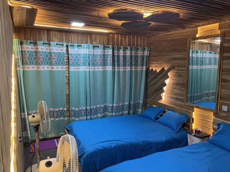 Bright cozy bedroom with wooden walls, blue bedding, patterned curtains, and warm lighting for a relaxing stay.